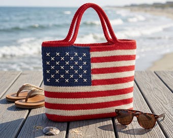 May include: A red, white, and blue crocheted tote bag with the American flag design. The bag has red handles and is sitting on a wooden deck near the beach. Brown sunglasses and sandals are also on the deck.