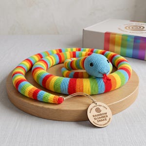 Op de afbeelding: Een gehaakt slangen-speeltje met een regenboog-gestreept lichaam, een lichtblauwe kop, zwarte ogen en een rode tong. Een rond houten label aan de staart bevat de tekst "HAND-CRAFTED TOYS RAINBOW SLUMBER SNAKE PREMIUM AMIGURUMI".