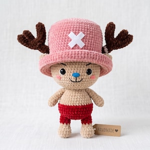 May include: A crocheted plush figure featuring a tan body, red shorts, and a pink hat with a white cross emblem. The figure has dark brown antlers, black eyes, a blue nose, and pink cheeks. A small brown tag labelled HANDMADE is attached.