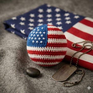 May include: A crocheted ball in the design of the American flag, with red and white stripes and a blue field with white stars. A military dog tag and a small stone are next to the ball. A folded American flag is in the background.