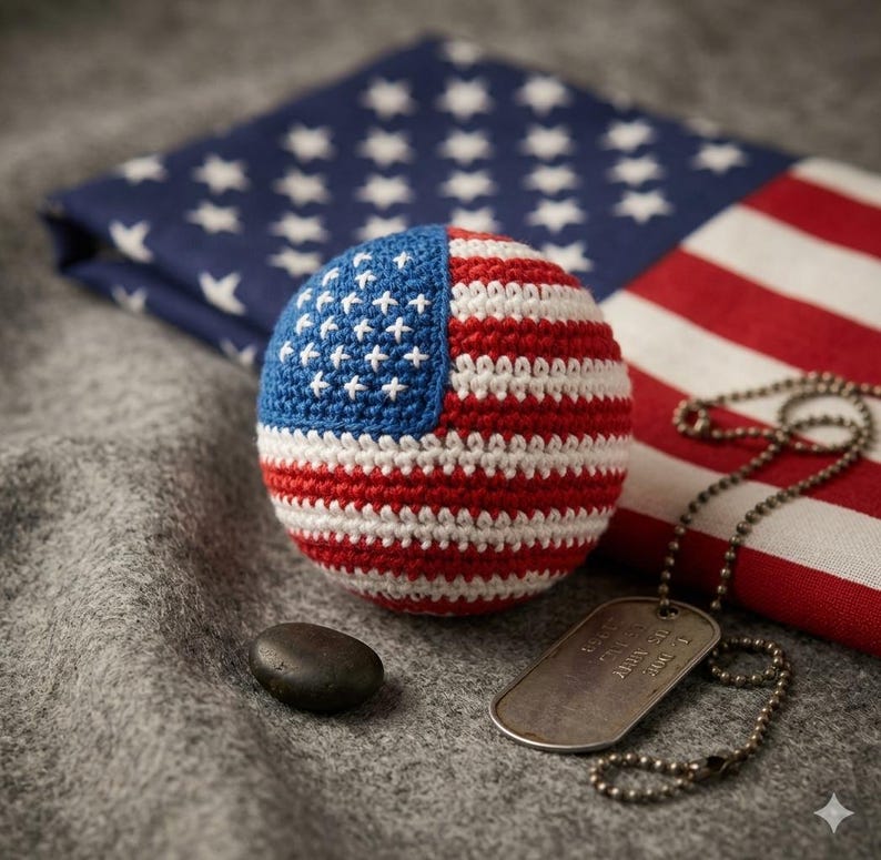 May include: A crocheted ball in the design of the American flag, with red and white stripes and a blue field with white stars. A military dog tag and a small stone are next to the ball. A folded American flag is in the background.