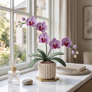 May include: A crocheted orchid plant with light purple petals and dark magenta centers, featuring long green leaves and small round buds. The plant is potted in a textured, cream-colored crocheted container with a brown top, resting on a gold hexagonal base.