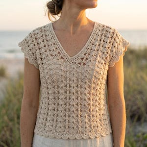 May include: A beige crochet top featuring a V-neckline and short cap sleeves. The garment is constructed with a repeating fan-like stitch pattern throughout the body and sleeves, finished with a scalloped edge along the hem and sleeve openings.