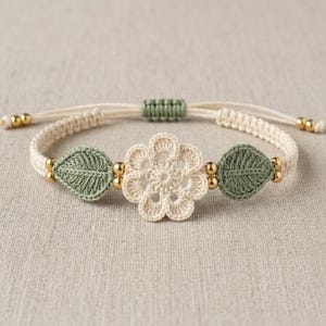 May include: A macrame bracelet featuring a central cream-colored crocheted flower flanked by two sage green crocheted leaves. The band is made of cream cord with a sage green adjustable sliding knot and small gold-toned metallic beads.