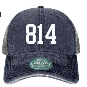 May include: A dark blue distressed trucker hat featuring the white number 814 printed on the front. The cap has a mesh back and a curved brim with a green circular sticker that reads Legacy Dashboard Trucker and #liveforlegacy on the front.