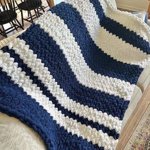 Op de afbeelding: Een grof gebreide plaid met een herhalend golvend patroon van dik, zacht marineblauw en wit garen. De deken is geconstrueerd met grote, pluche lussen van garen, wat zorgt voor een sterk getextureerd, tactiel oppervlak over het hele item.