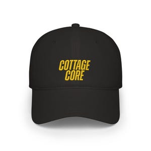 Könnte beinhalten: Schwarze Baseballkappe mit gebogenem Schirm. Auf der Kappe ist der Text "COTTAGE CORE" in Gelb aufgestickt. Der Hut hat ein klassisches Sechs-Panel-Design.