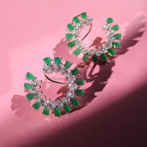 May include: A pair of silver-toned earrings with a curved design. Each earring features a series of green teardrop-shaped stones and clear, sparkling stones. The earrings are set against a pink background.