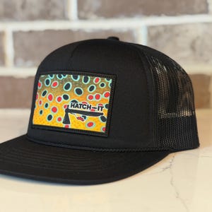 May include: A black trucker hat featuring a rectangular patch on the front. The patch displays a colorful brown trout skin pattern with red and black spots and the text HATCH-IT in black letters above a stylized black fishing fly graphic.