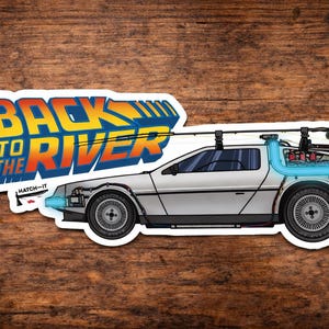 May include: A die-cut sticker featuring a silver sports car with a fishing rod mounted on the roof and a glowing blue tube attached to the rear. The text "BACK TO THE RIVER" is printed in a bold, retro-style font above the car, with "HATCH-IT" below.
