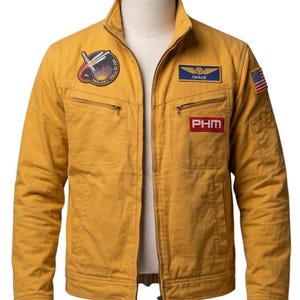 May include: A mustard yellow jacket with a stand-up collar and a full-length front zipper. The chest features a circular mission patch, a blue name tag with the text GRACE, and a red rectangular patch with the text PHM. A small American flag patch is on the sleeve.