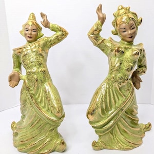 May include: Two ceramic figurines depicting dancers in dynamic poses. Both figures wear lime green outfits with gold-toned accents and textured finishes. The figures feature elaborate headpieces and are captured in mid-motion with arms raised.