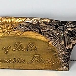 May include: A gold-toned metal souvenir scoop featuring an embossed floral pattern along the handle and top edge. The center is engraved with the text "Souvenir of Dells of the Wisconsin River" in a cursive script, set against a smooth surface.