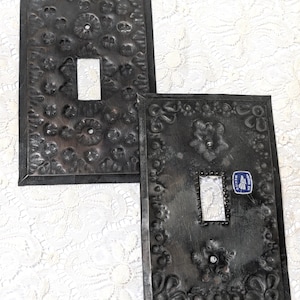 May include: Two rectangular dark metal light switch plates featuring embossed floral patterns and a central rectangular cutout. One plate displays a small blue and white sticker with the text Hecho en Mexico near the center opening.