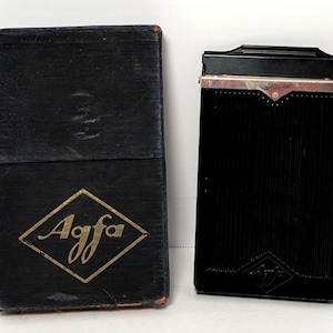 May include: A vintage black Agfa film plate holder and its matching dark blue fabric-covered storage box. The box features the gold Agfa logo in a diamond shape on the front. The film holder is made of black material with a metallic top bar.