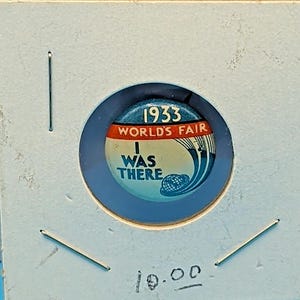 May include: A circular pinback button featuring a blue, white, and red design with the text 1933 WORLD'S FAIR I WAS THERE. The button displays a graphic of a globe and curved lines, encased in a square cardboard holder with the number 10.00 written.