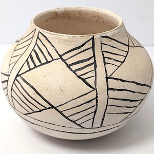 May include: A round, off-white ceramic pot featuring a wide, open mouth and a matte, textured surface. The exterior is decorated with hand-painted black geometric patterns consisting of intersecting lines and triangles that wrap around the body.