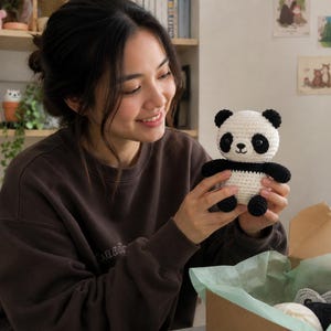 Könnte beinhalten: Ein handgehäkelter Panda-Bär mit schwarzen und weißen Details. Der Panda hat schwarze Augen, Ohren und Gliedmaßen und einen weißen Körper. Der Panda wird in der Nähe einer offenen Pappschachtel mit anderen Garnartikeln gehalten.