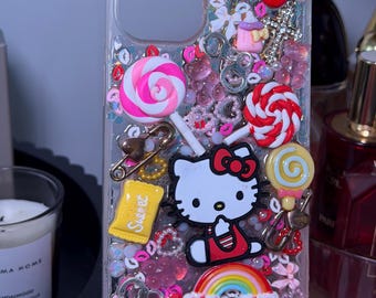 May include: Clear phone case decorated with a variety of charms, including a Hello Kitty figure, lollipops, bows, and a rainbow. The case has a glittery background and a variety of pink, red, and yellow accents. The word "Sweet" is visible on a yellow candy wrapper.