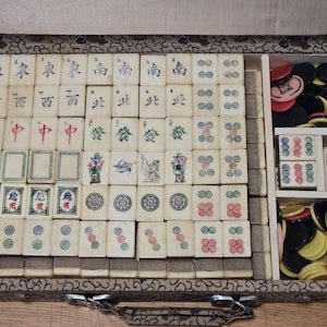 Op de afbeelding: Een vintage Mahjong-spel in een met patroonstof beklede koffer, met gebroken witte tegels voorzien van gegraveerde Chinese karakters, bloemmotieven en cirkelvormige patronen. De set bevat ronde speelfiches in zwart, rood en geel plastic.