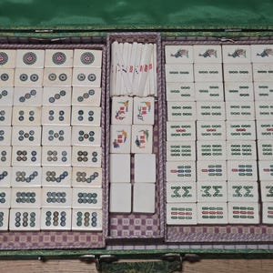 May include: A mahjong set housed in a green case, featuring rectangular tiles with various circular, numeric, and character designs in red, blue, and green ink. The set includes a central compartment containing thin white scoring sticks and blank tiles.