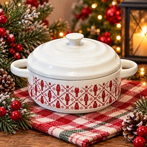 May include: A white ceramic lidded casserole dish featuring a repeating red pattern of nutcracker figures and geometric diamond shapes. The round pot has two side handles and a matching lid with a central knob, finished in a glossy glaze.