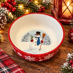 May include: A round ceramic bowl with a bright red rim and a white interior featuring a painted snowman wearing a black top hat and blue scarf, holding a broom. The exterior of the bowl is red with white snowflake patterns around the sides.