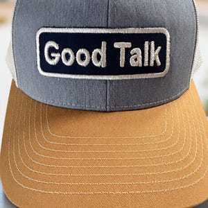 May include: A gray and tan baseball cap featuring a rectangular patch on the front panel with a dark blue background and white embroidered text that reads Good Talk. The cap has a tan brim with white stitching and a mesh back panel.