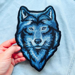 Puede incluir: Un parche bordado que presenta la cabeza de un lobo en tonos azules, blancos y negros. El diseño muestra costuras detalladas para crear la textura del pelaje, orejas puntiagudas y ojos enfocados, todo delineado con un borde negro grueso.