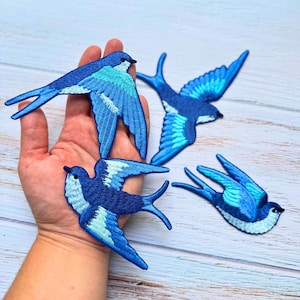May include: Four embroidered swallow patches in varying shades of blue, including navy, royal blue, and light cyan. Each bird is depicted in flight with wings spread, featuring detailed stitching that defines the feathers and body shape.