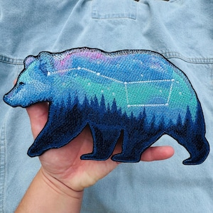 May include: An embroidered bear-shaped patch featuring a dark blue forest silhouette at the bottom, transitioning into a vibrant blue and purple night sky with a white stitched constellation pattern across the upper back of the bear figure.