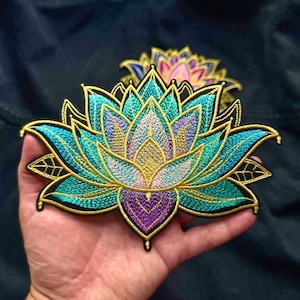 May include: An embroidered lotus flower patch featuring vibrant teal, purple, and gold thread work. The design showcases intricate petal detailing with a bold black border, highlighting a symmetrical floral pattern with layered stitching throughout.