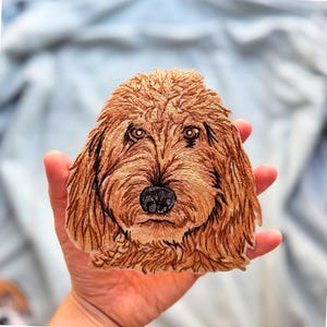 May include: An embroidered patch featuring the detailed head of a golden-brown dog with shaggy fur, dark eyes, and a black nose. The stitching captures the texture of the coat with varied shades of tan and brown thread on a fabric base.