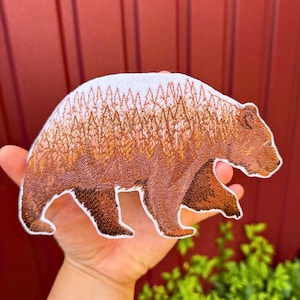 Può includere: Una toppa ricamata a forma di orso che cammina. Il corpo dell'orso presenta un dettagliato motivo paesaggistico con strati di pini marroni e arancioni su sfondo bianco, creando un effetto di doppia esposizione sulla sagoma dell'animale.