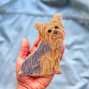 May include: An embroidered patch shaped like a Yorkshire Terrier dog. The design features detailed stitching in shades of golden tan and dark grey, capturing the texture of the dog's fur, with dark eyes and a small black nose on the face.