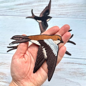 Pode incluir: Patches bordados de andorinhas com detalhes em fio castanho, preto e branco. Os designs das aves mostram as andorinhas em voo com as asas estendidas e caudas bifurcadas. Estes patches de tecido são feitos com costuras detalhadas.