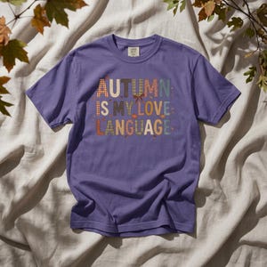 May include: A purple short-sleeve t-shirt featuring the text "AUTUMN IS MY LOVE LANGUAGE" in a multi-colored, patterned block font. The letters include various plaid, checkered, and solid autumn-toned designs, accented with small leaf and bow icons.