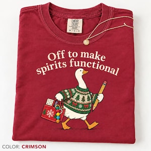 May include: A crimson t-shirt featuring a graphic of a white goose wearing a green patterned sweater, holding a yellow pencil and a red tote bag with a snowflake design. The text reads Off to make spirits functional. A gold necklace rests on top.