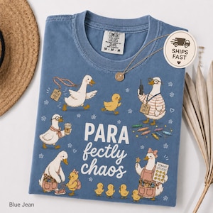 May include: A blue jean colored t-shirt featuring a graphic print of multiple ducks and ducklings. The text reads PARA fectly chaos. The ducks are depicted in various activities, including wearing lanyards labeled PARA PRO, holding a coffee cup, and carrying a reward sticker chart. The shirt has a Comfort Colors brand label at the neck.