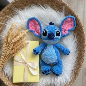 Puede incluir: Un peluche de ganchillo azul con orejas grandes con detalles interiores rosas, ojos negros y nariz azul oscuro. El personaje tiene una mancha azul claro en el vientre y detalles azul oscuro en manos y pies, hecho con hilo suave y texturizado.