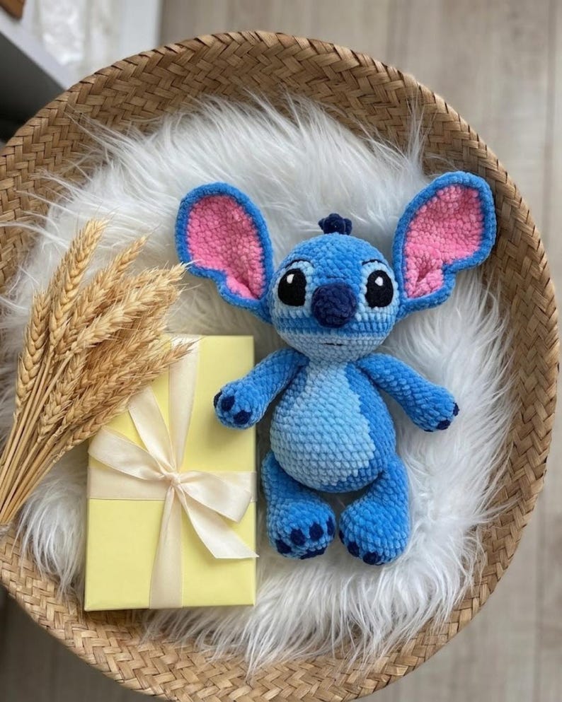 Puede incluir: Un peluche de ganchillo azul con orejas grandes con detalles interiores rosas, ojos negros y nariz azul oscuro. El personaje tiene una mancha azul claro en el vientre y detalles azul oscuro en manos y pies, hecho con hilo suave y texturizado.