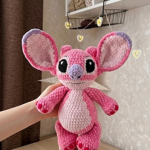 Puede incluir: Un peluche de ganchillo de un personaje alien&iacute;gena rosa con orejas grandes, una mancha rosa claro en el vientre y una nariz morada. La figura presenta ojos bordados en negro, una peque&ntilde;a boca cosida y detalles en rosa oscuro en los pies y las orejas.