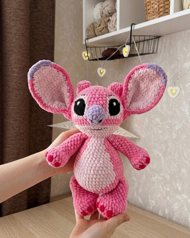 Puede incluir: Un peluche de ganchillo de un personaje alien&iacute;gena rosa con orejas grandes, una mancha rosa claro en el vientre y una nariz morada. La figura presenta ojos bordados en negro, una peque&ntilde;a boca cosida y detalles en rosa oscuro en los pies y las orejas.