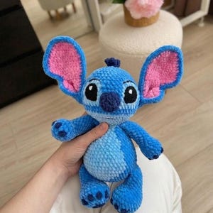 Puede incluir: Un peluche de ganchillo azul con orejas grandes y detalles interiores rosas, cabeza redonda con ojos bordados negros, nariz azul oscuro y una mancha azul claro en el vientre. El personaje tiene cuatro extremidades con almohadillas oscuras.