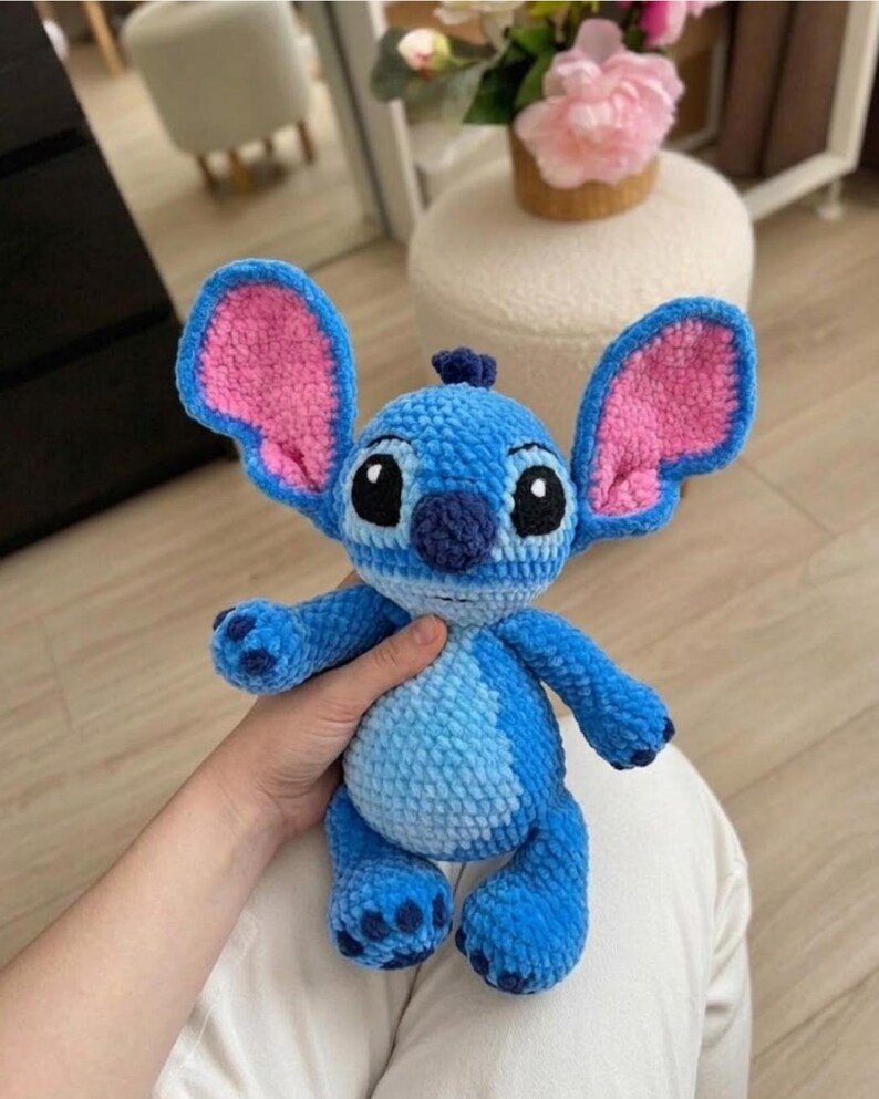 Puede incluir: Un peluche de ganchillo azul con orejas grandes y detalles interiores rosas, cabeza redonda con ojos bordados negros, nariz azul oscuro y una mancha azul claro en el vientre. El personaje tiene cuatro extremidades con almohadillas oscuras.
