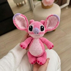 Puede incluir: Un peluche tejido a mano en crochet que representa un personaje alien&iacute;gena rosa con orejas grandes, una mancha rosa claro en el vientre, nariz morada y ojos negros. La construcci&oacute;n con hilo texturizado le da un aspecto suave y detallado.