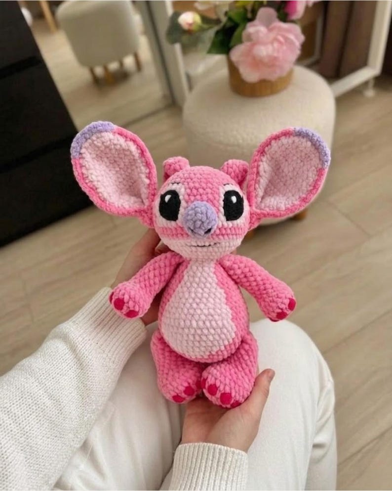 Puede incluir: Un peluche tejido a mano en crochet que representa un personaje alien&iacute;gena rosa con orejas grandes, una mancha rosa claro en el vientre, nariz morada y ojos negros. La construcci&oacute;n con hilo texturizado le da un aspecto suave y detallado.