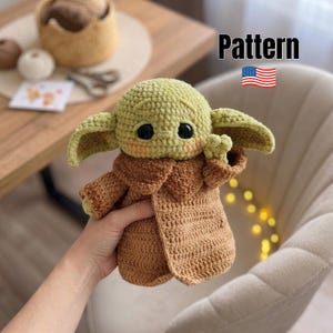 Op de afbeelding: Een gehaakt knuffeltje van een klein groen wezen met grote zwarte ogen, puntige oren en roze wangen, gekleed in een bruine jas met textuur. De afbeelding bevat de tekst Pattern en een klein icoon van een Amerikaanse vlag.
