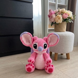 Puede incluir: Un peluche de ganchillo hecho a mano que representa un personaje alien&iacute;gena rosa y rosa claro con orejas grandes, nariz morada y ojos negros. La figura tiene un cuerpo redondeado, brazos cortos y pies con almohadillas circulares rosa oscuro.