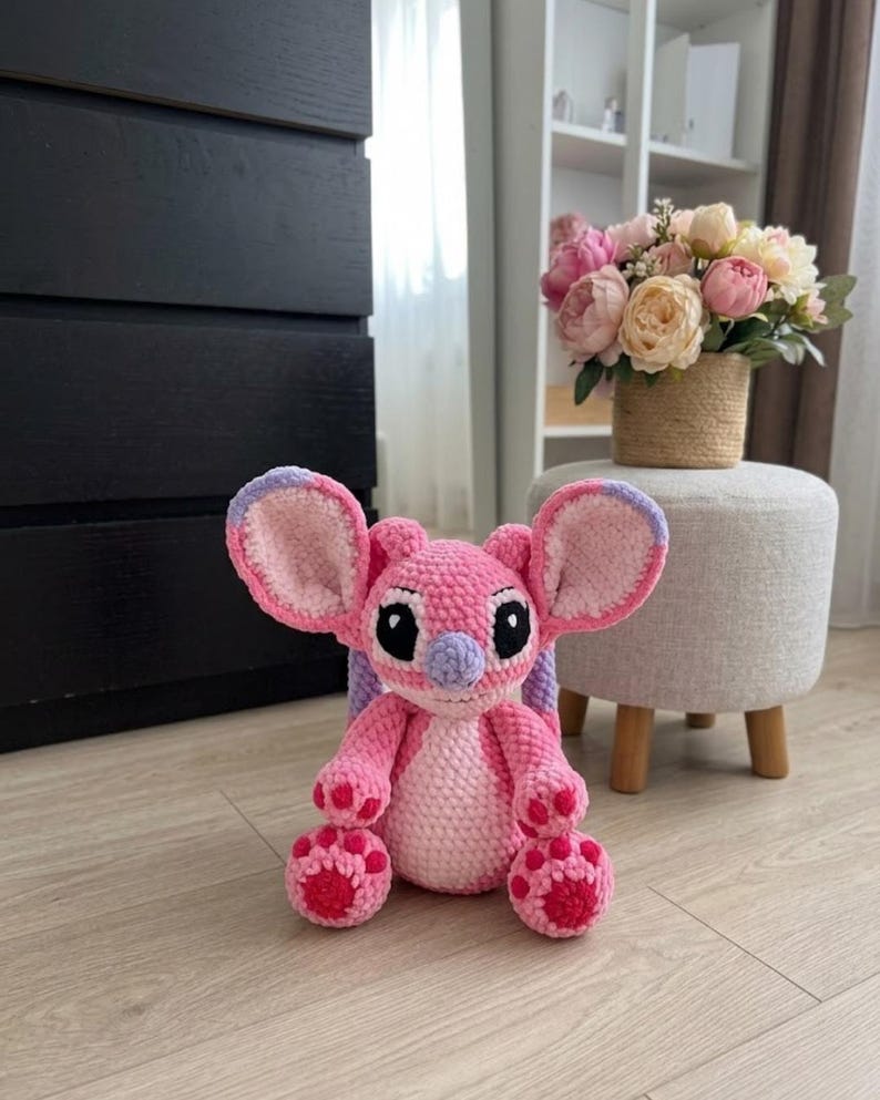 Puede incluir: Un peluche de ganchillo hecho a mano que representa un personaje alien&iacute;gena rosa y rosa claro con orejas grandes, nariz morada y ojos negros. La figura tiene un cuerpo redondeado, brazos cortos y pies con almohadillas circulares rosa oscuro.